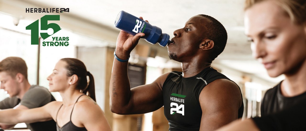 People using exercise equipment in a gym; one person drinks from a sports bottle while others exercise in the background.