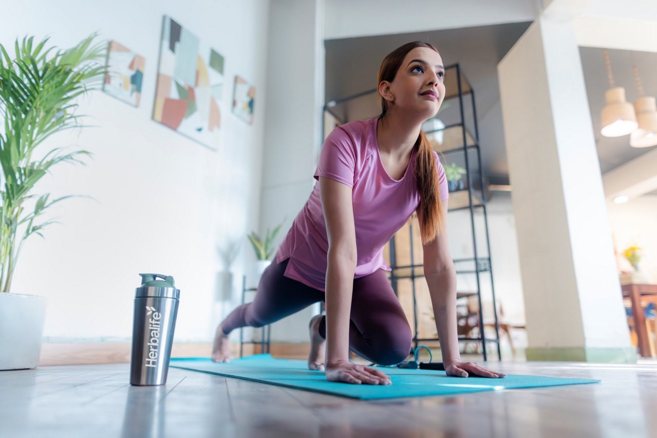 Woman performing yoga pose on a blue mat at home with a Herbalife bottle nearby, wearing pink top and purple leggings in a bright modern living space.