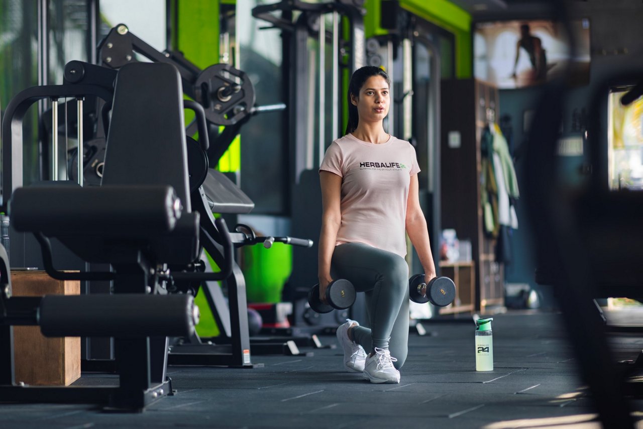 Woman in Herbalife t-shirt performing lunges with dumbbells in a modern gym with a Herbalife H24 bottle nearby.