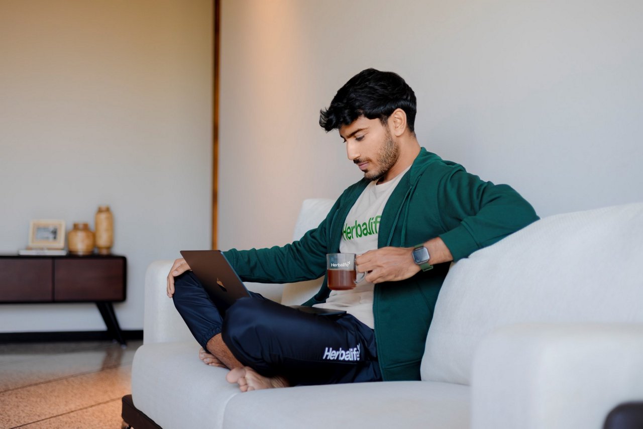 Man sitting on white sofa working on laptop while drinking from a Herbalife cup, wearing a green hoodie over a Herbalife t-shirt and black Herbalife sweatpants.