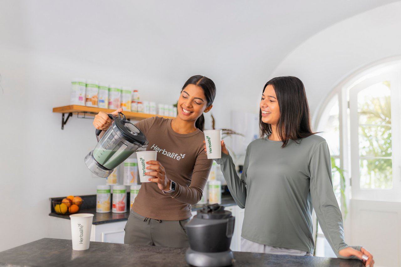 Two women preparing a smoothie using a blender, smiling and enjoying in a kitchen setting.