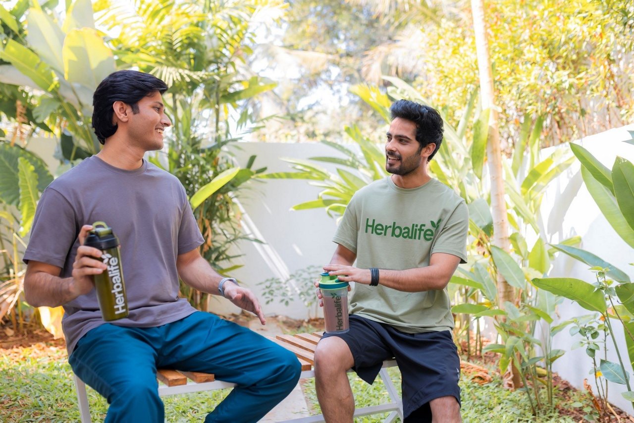 Two men sitting on a bench in a garden setting, holding Herbalife products and having a conversation surrounded by tropical plants.