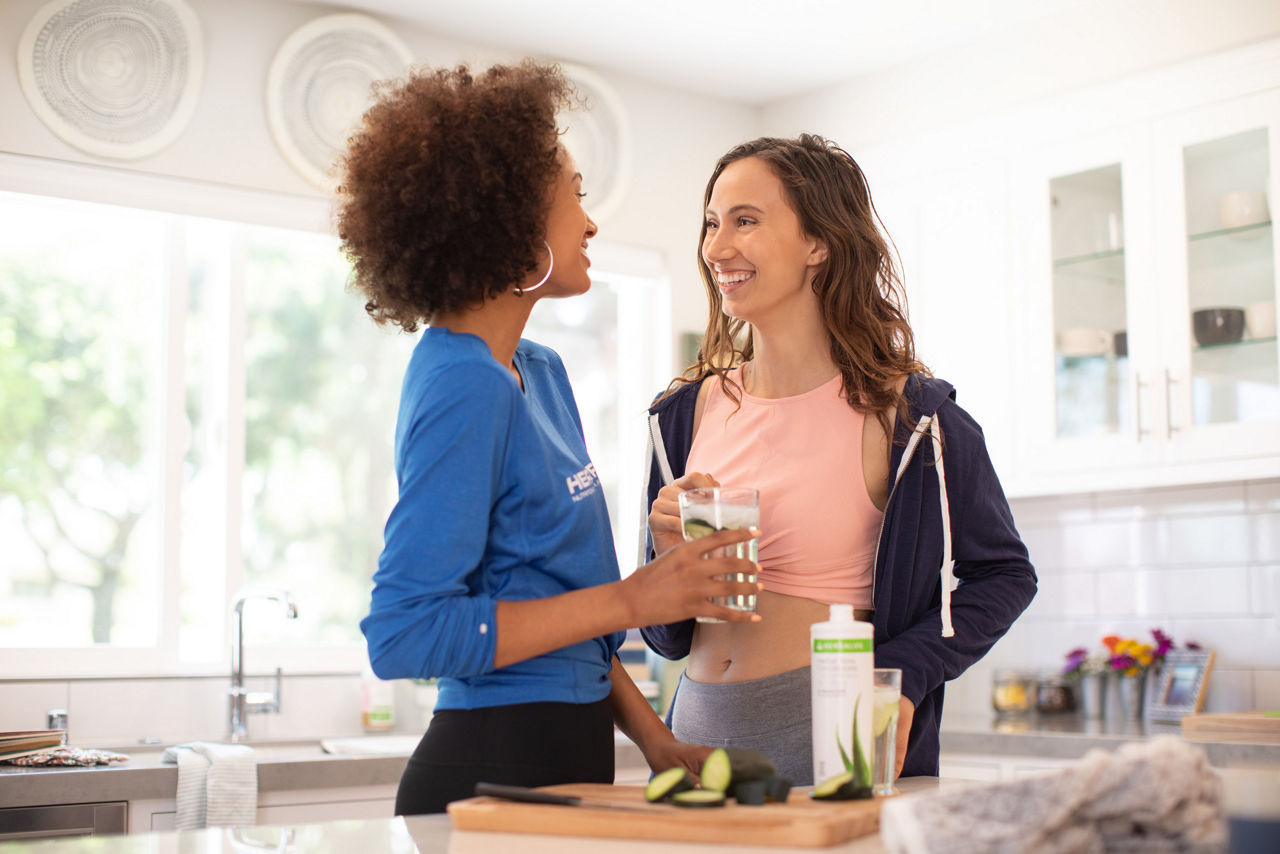 Two women in kitchen enjoying a glass of an Herbal Aloe beverage