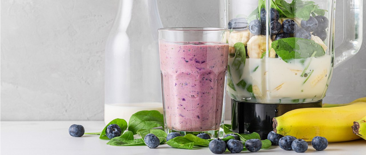 A glass of blueberry, banana and spinach protein shake with fresh ingredients in a blender