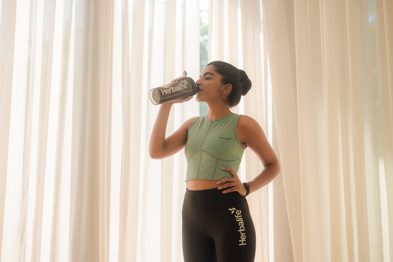 Asian woman in activewear drinking from Herbalife-branded bottle, bright interior background.