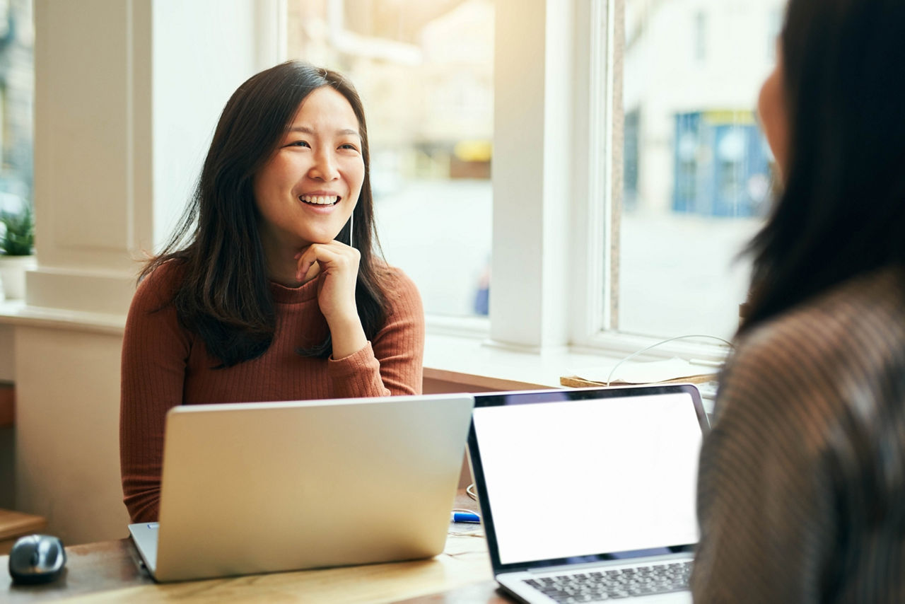 Two young women talk while working on their laptops