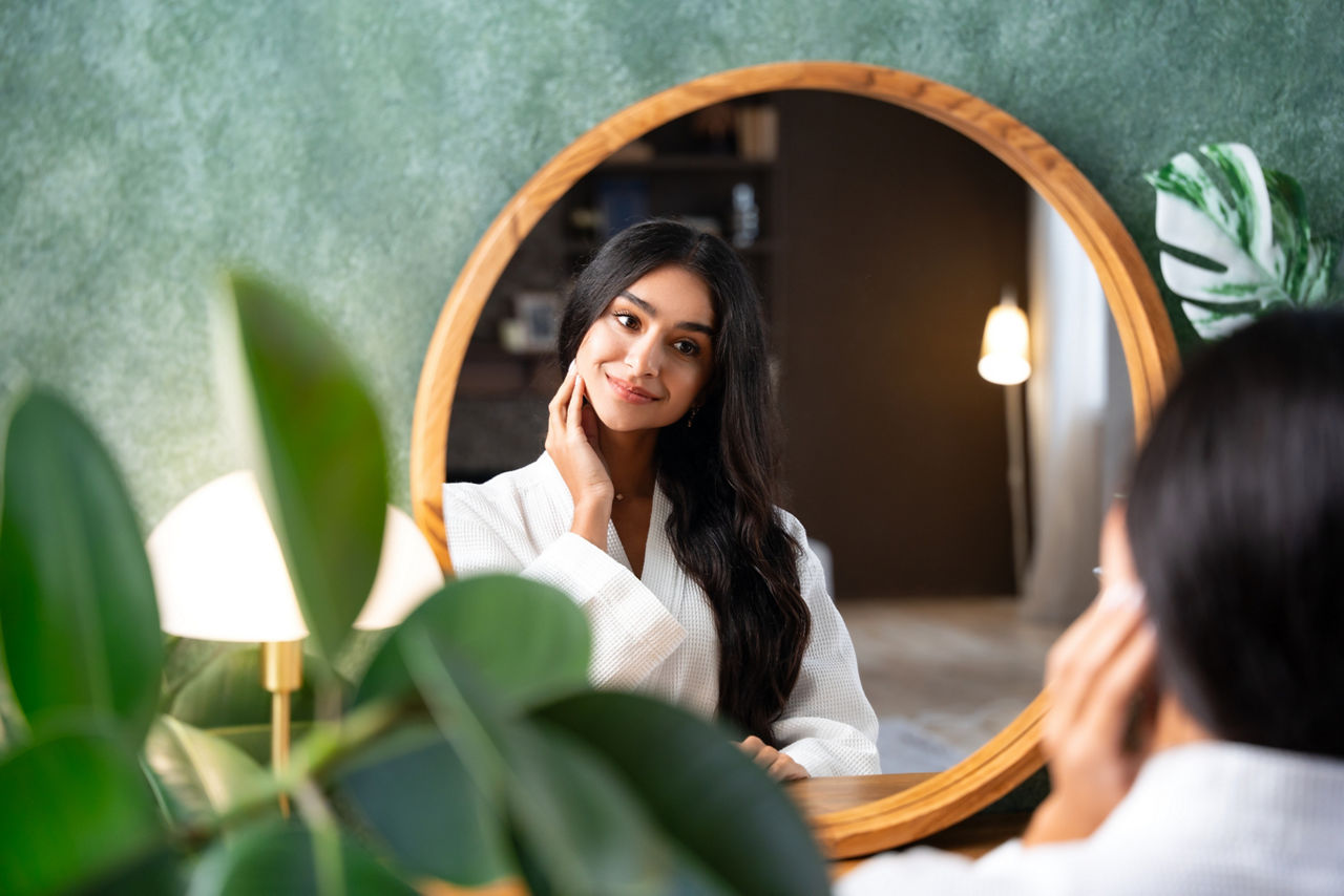 Hair care and self-care with beautiful Indian woman looking in mirror touching her healthy long hair sitting at the dressing table. Interior of the room is in green colors and with an abundance of green plants.